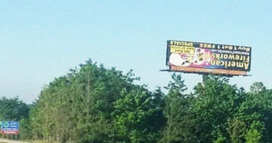 A billboard advertising American Fireworks stands above green trees beside a highway under a clear blue sky. The sign is partially readable and slightly tilted.