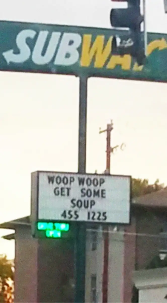 A blurry photo of a Subway restaurant sign reads, "WOOP WOOP GET SOME SOUP 455 1225" beneath the Subway logo. There is a traffic light and part of a building in the background.