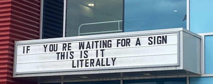 A marquee sign on a building reads, "IF YOU'RE WAITING FOR A SIGN THIS IS IT LITERALLY." The background includes blue windows and red siding.