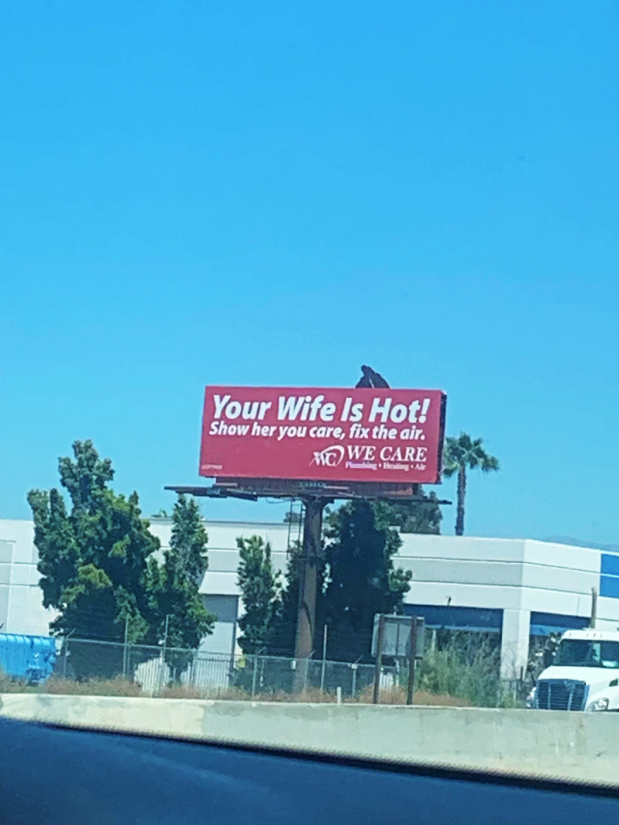 A red billboard reads, "Your Wife Is Hot! Show her you care, fix the air. We Care." It is located above a building with trees nearby, under a clear blue sky.