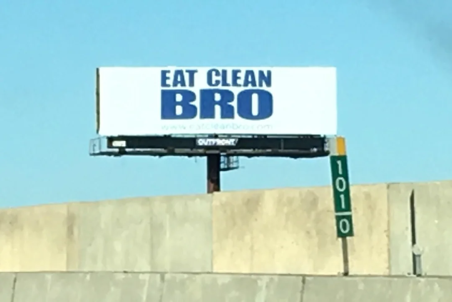 A roadside billboard with the words "EAT CLEAN BRO" in bold blue letters on a white background, positioned above a concrete barrier and next to a green highway marker labeled "101 0".
