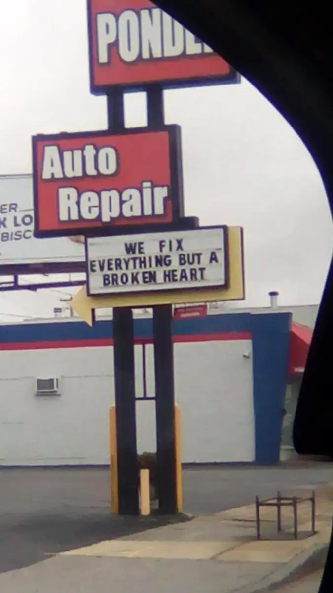 A sign outside an auto repair shop reads, "WE FIX EVERYTHING BUT A BROKEN HEART," with the shop and a yellow arrow visible in the background.