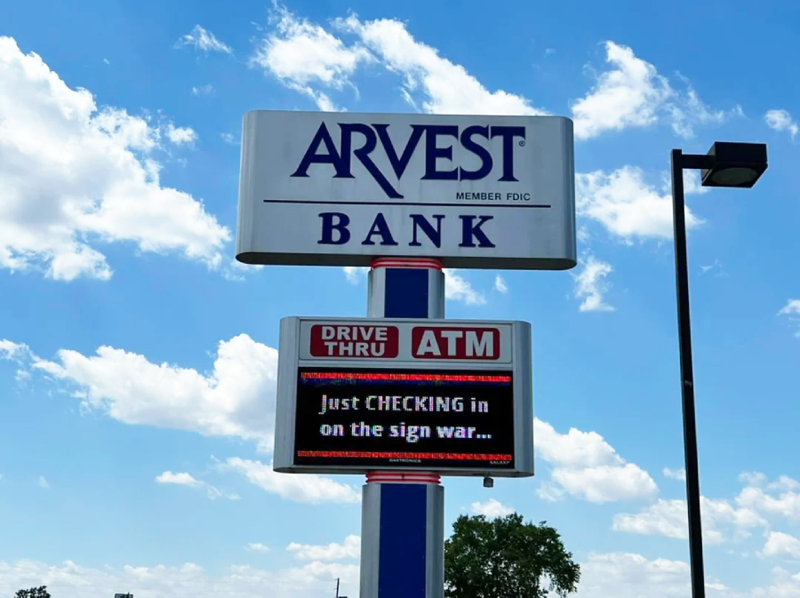 A sign outside Arvest Bank displays “Just CHECKING in on the sign war…” beneath a “DRIVE THRU ATM” label, against a blue sky with scattered clouds.