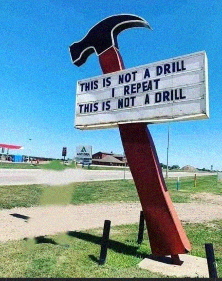 A large red hammer sculpture holds a roadside sign that reads, "THIS IS NOT A DRILL I REPEAT THIS IS NOT A DRILL" against a clear blue sky with buildings in the background.