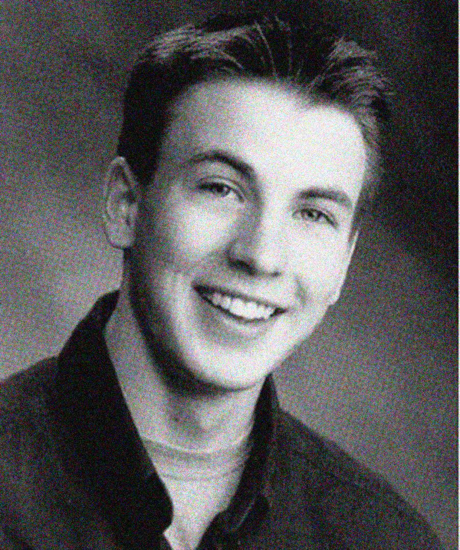 A black-and-white portrait of a smiling young man with short, neatly combed hair, wearing a collared shirt over a T-shirt, looking directly at the camera against a blurred background.