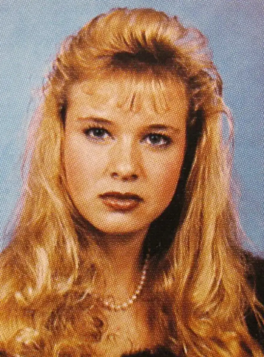 A woman with long, wavy blonde hair and blue eyes is wearing a pearl necklace. She has a serious expression and is posed in front of a blue background.