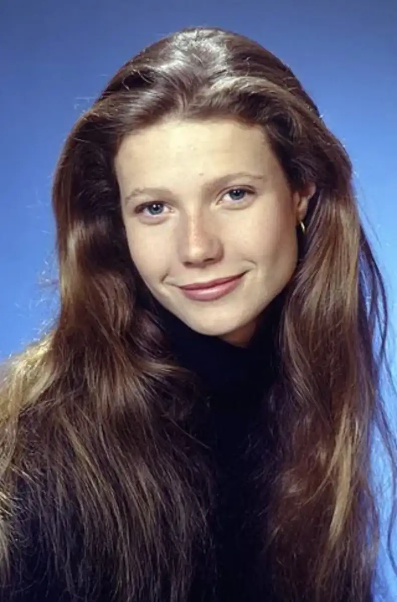 A young woman with long, straight brown hair and blue eyes smiles softly. She is wearing a black turtleneck and small hoop earrings, with a plain blue background behind her.