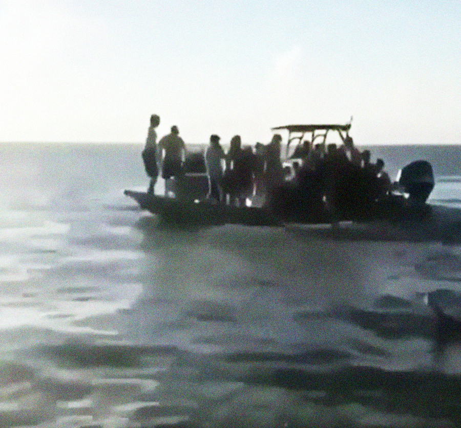 A group of people stand closely together on a small boat in open water under a bright sky. The water appears calm, and the scene is slightly blurry.