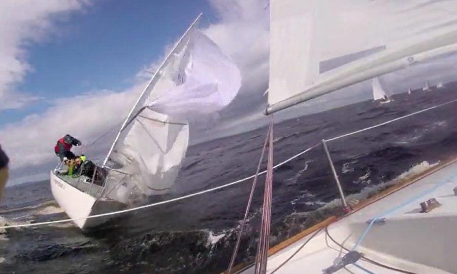 A sailboat leans sharply to the side in choppy water, its sails tangled and flapping, while crew members work on deck. The photo is taken from another nearby sailboat. The sky is partly cloudy.