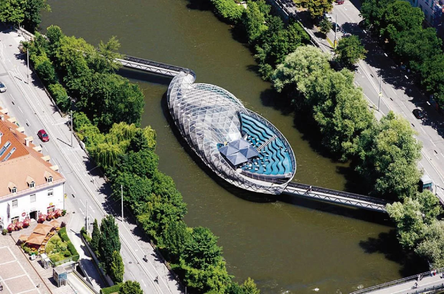 Aerial view of an oval-shaped, futuristic floating structure with a glass roof and blue seating on a river, connected to both riverbanks by footbridges and surrounded by trees and city streets.