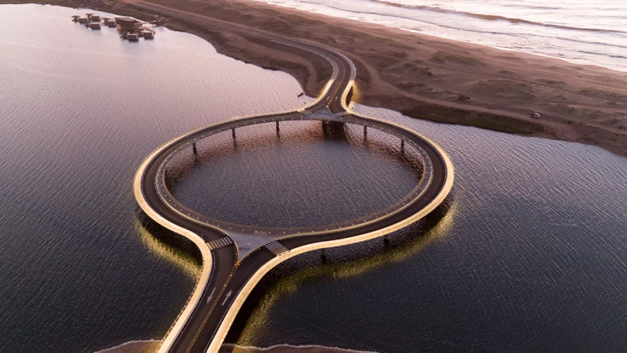 Aerial view of a circular bridge over calm water extending from a sandy shoreline, with gentle waves visible in the background and soft lighting illuminating the bridge at dusk.