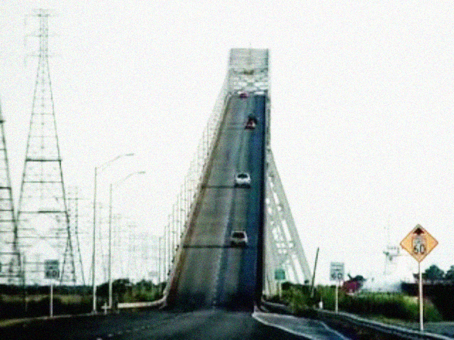 A steep, elevated bridge with several cars driving up and down its incline, flanked by power line towers. A yellow caution sign and speed limit sign are visible on the right side of the road.