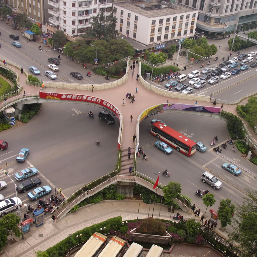 Aerial view of a busy intersection with a clover-shaped pedestrian overpass, cars, buses, and people crossing. Surrounding buildings, greenery, and traffic can be seen.