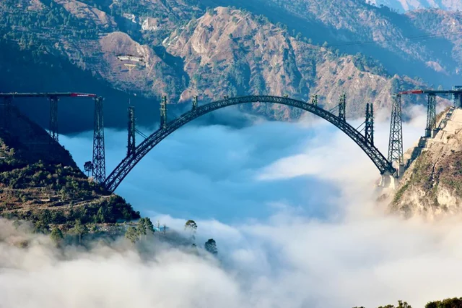A large steel arch bridge spans between two mountains above a valley filled with dense clouds and mist, with rugged hills and sparse vegetation visible in the background.