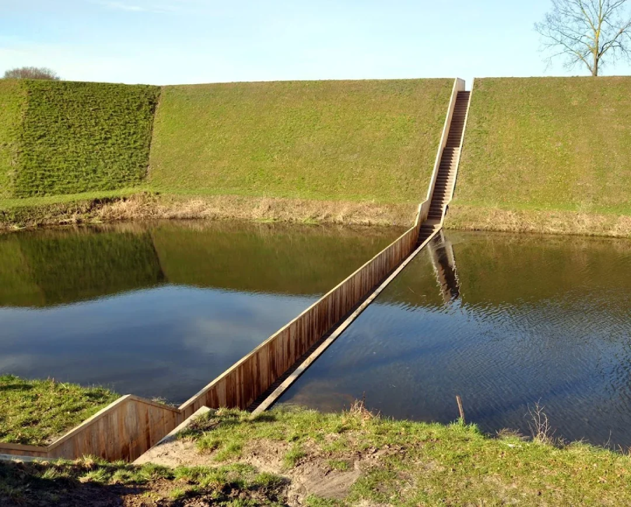 A sunken wooden walkway, known as the Moses Bridge, cuts through a grassy embankment and a reflective moat, creating the visual illusion of walking through water.
