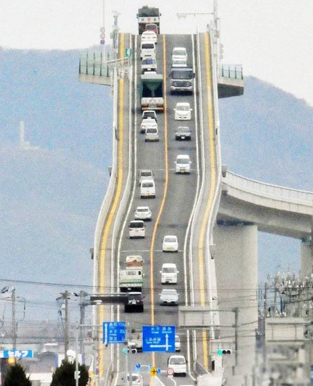 A steep, elevated bridge with cars driving up and down its wavy, roller coaster-like incline against a hazy background of hills and power lines.