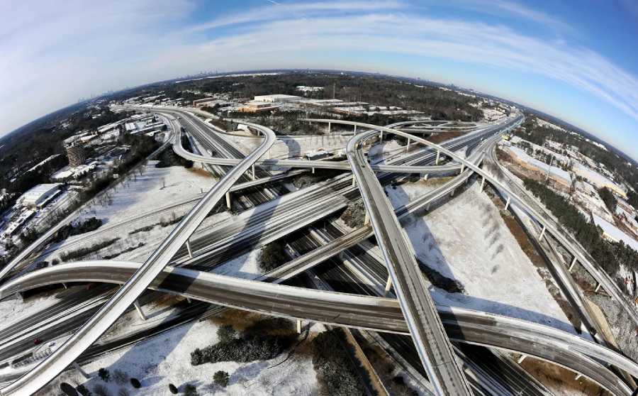 Aerial view of a large, complex highway interchange with multiple overlapping roads and ramps, surrounded by snow-covered ground and sparse trees on a clear day.