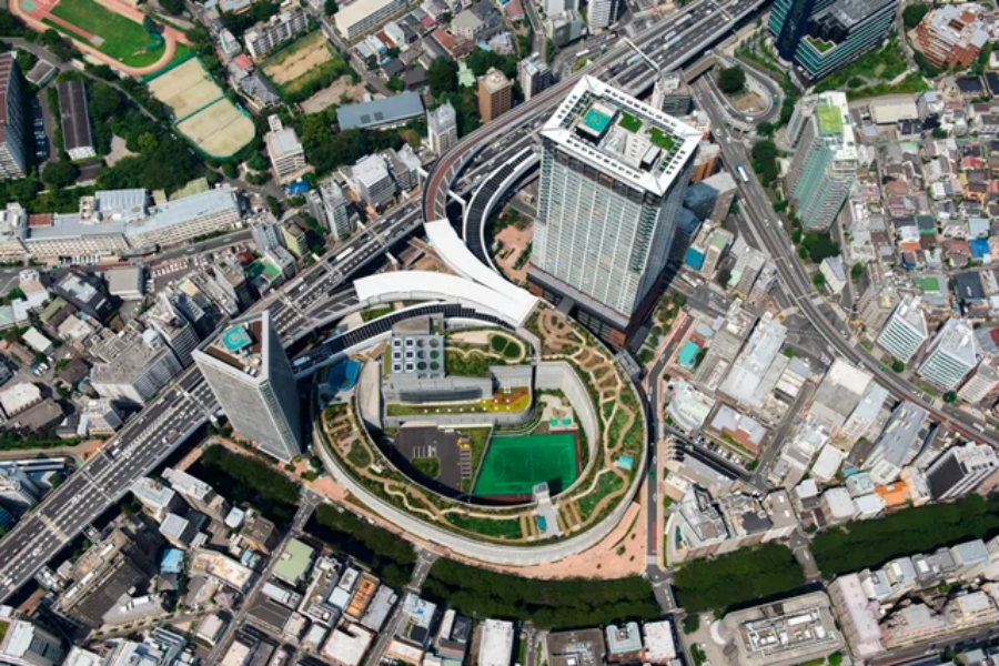 Aerial view of a modern cityscape featuring tall buildings, curved roads, a rooftop tennis court, and densely packed surrounding structures, with green spaces interspersed throughout.