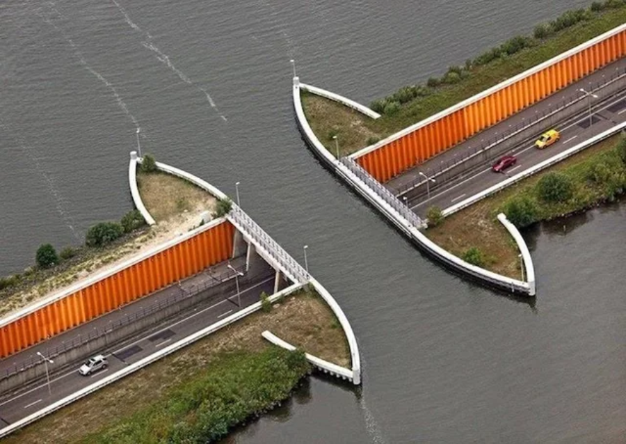 Aerial view of two roads passing under a waterway with orange walls, allowing boats to cross over the roads. Cars are driving on both roads, surrounded by green vegetation and water.