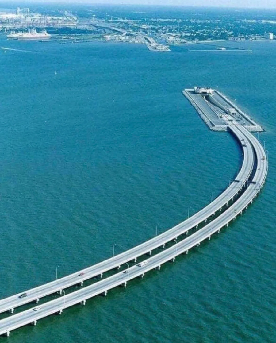 Aerial view of a curved bridge extending over blue water, with cars traveling on it and a city visible in the distant background. The bridge leads to a ramp that appears to end abruptly.
