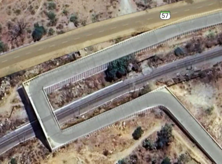 Aerial view of a road making a sharp zigzag turn over a railway track in a dry, rocky landscape. A road sign with the number 57 is visible on the upper road. Sparse vegetation is scattered around.