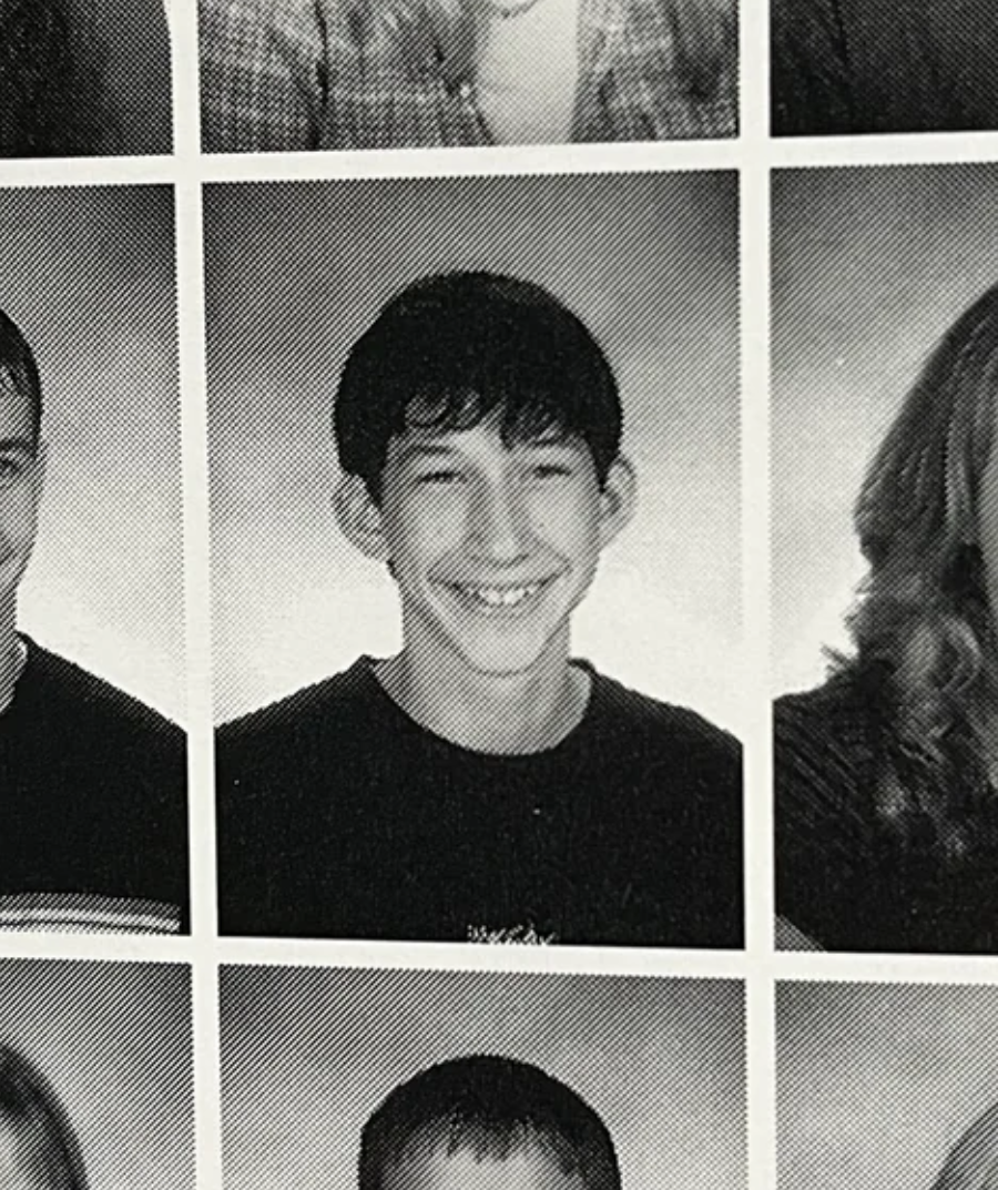 Black-and-white yearbook photo of a teenage boy with short, dark hair, smiling broadly. He is wearing a dark shirt and is centered in the frame, with partial views of other classmates surrounding him.