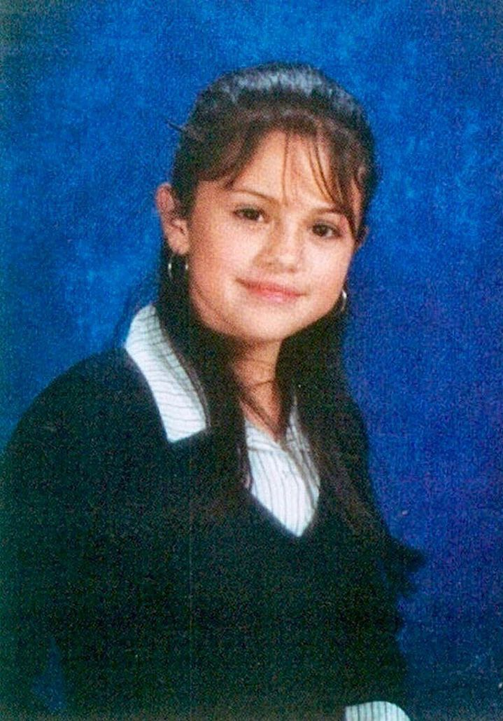 A young girl with long dark hair, wearing a striped collared shirt and a dark sweater, poses for a school portrait against a blue background. She is smiling slightly and looking at the camera.