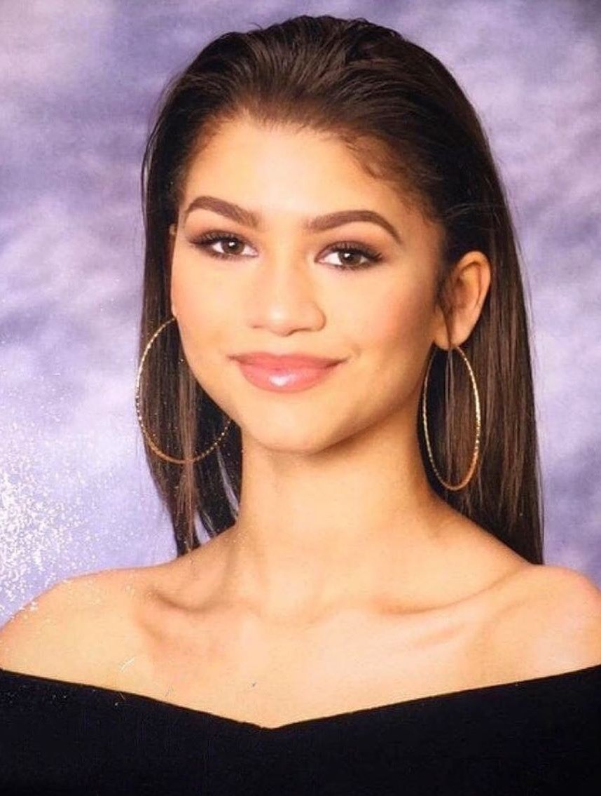 A young woman with long straight hair, wearing large hoop earrings and an off-the-shoulder black top, poses in front of a soft purple and white studio backdrop, smiling slightly.