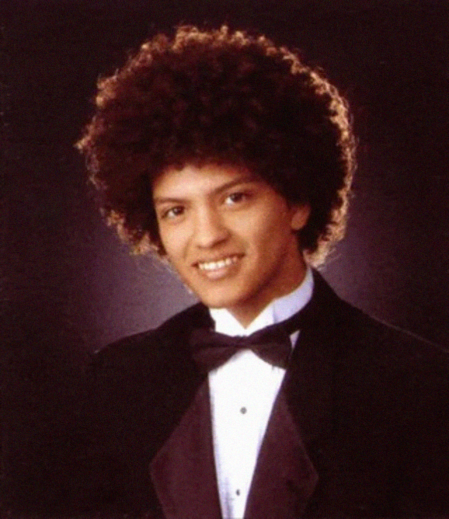 A young man with curly hair is smiling, wearing a black tuxedo jacket, white shirt, and black bow tie, posed against a dark, softly lit background.
