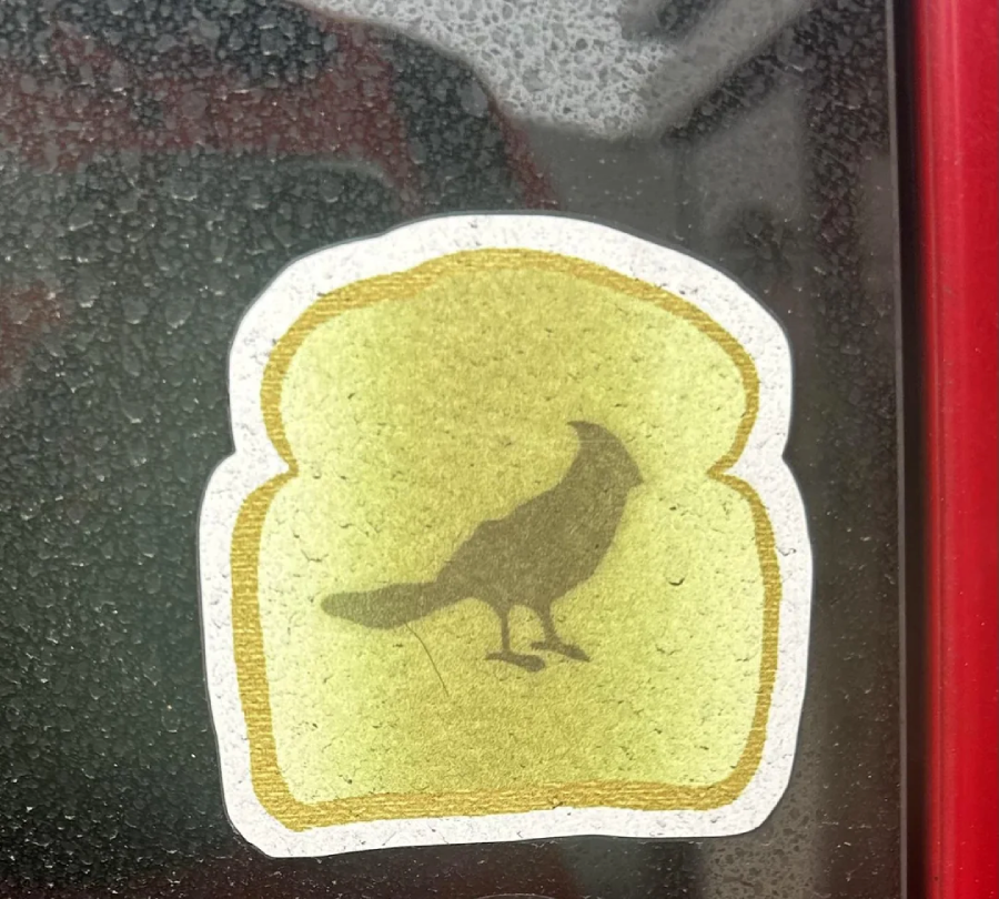 A sticker shaped like a slice of bread with a silhouette of a bird in the center is placed on a window covered in water droplets. A red border is visible on the right side of the image.