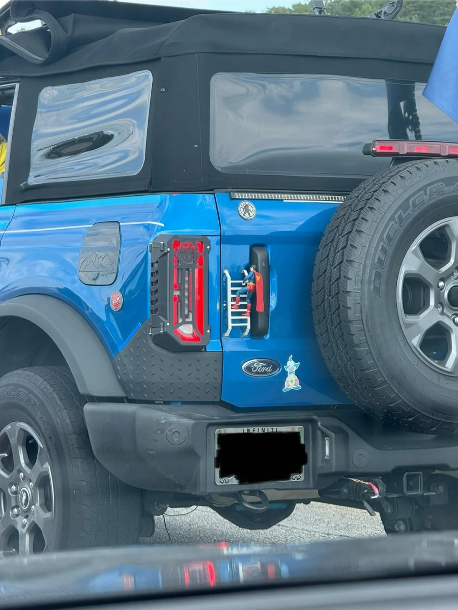A blue Ford Bronco with a black soft top, rear-mounted spare tire, and visible off-road accessories is parked outside. The vehicle has custom tail lights and a cartoon rabbit sticker near the Ford emblem.