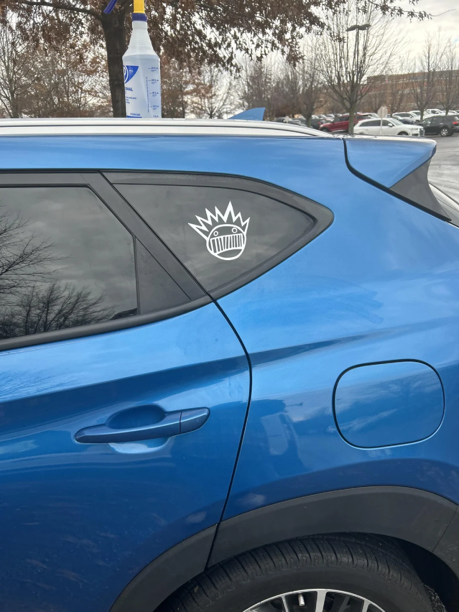 A blue car with a white sticker of a cartoonish face with a wide, jagged mouth and a spiky crown on the rear side window. Trees and other parked cars are visible in the background.