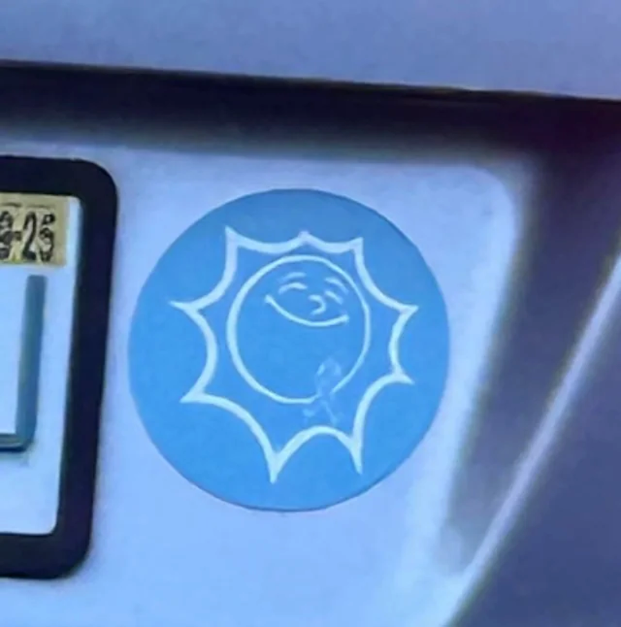 A blue circular sticker on a car shows a smiling sun with a mischievous face, appearing to urinate onto the car's surface. Part of a license plate is visible nearby.