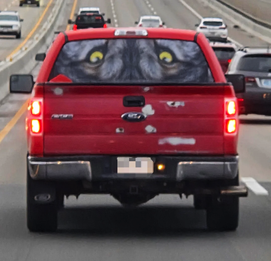A red pickup truck on a highway with a rear window decal showing large, yellow animal eyes staring outward, creating an illusion of a creature inside the truck.