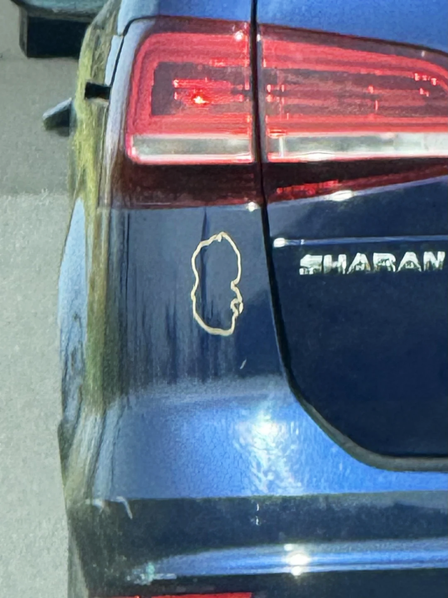 A close-up of the rear of a blue car shows a scratch or outline in the paint resembling the profile of a face or head near the model name "SHARAN" on the trunk.