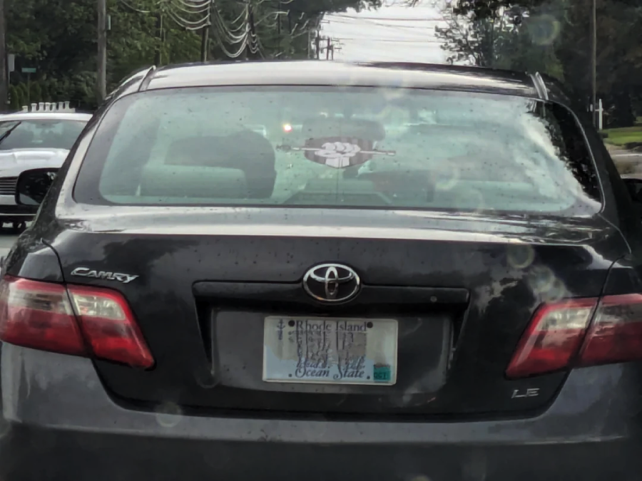 A black Toyota Camry with a Rhode Island license plate is seen from behind on a rainy day. A sticker with a raised fist graphic is displayed on the rear window.