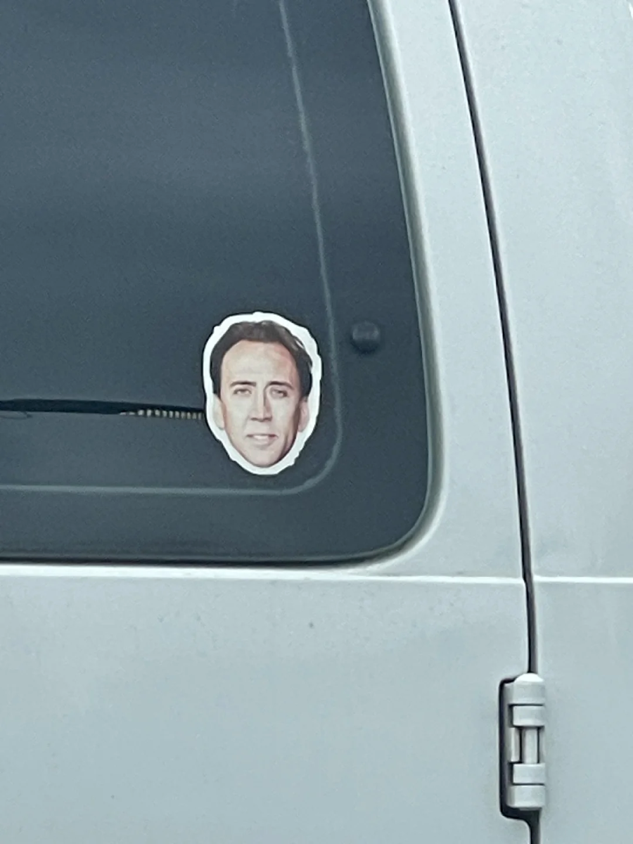 A sticker with a man's face is placed on the tinted window of a white vehicle near the door frame.