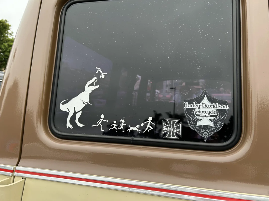 A car window decal shows a T. rex eating stick figure family members, with others fleeing. Nearby are Harley-Davidson and Iron Cross logo stickers. The vehicle is brown and beige.