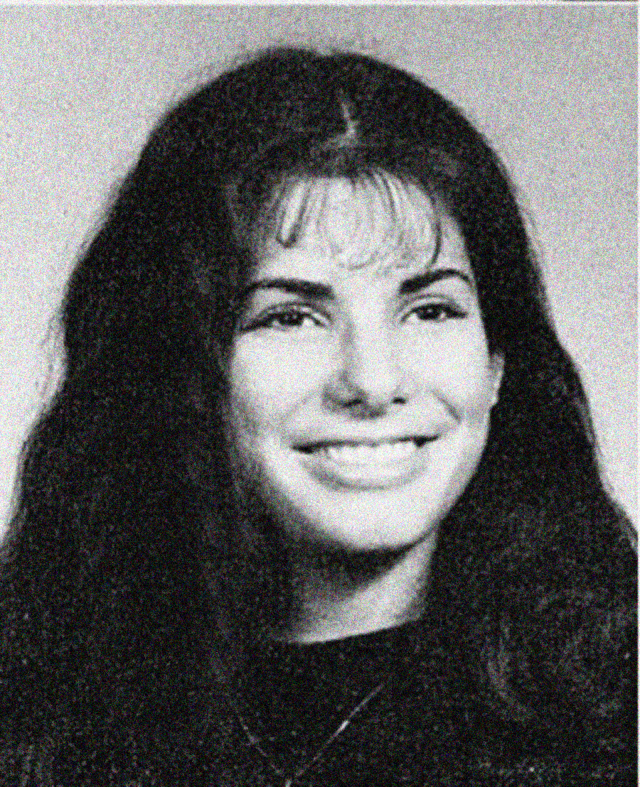 Black and white yearbook-style photo of a young woman with long dark hair and bangs, smiling at the camera. She is wearing a dark top and a necklace, and the background is plain.