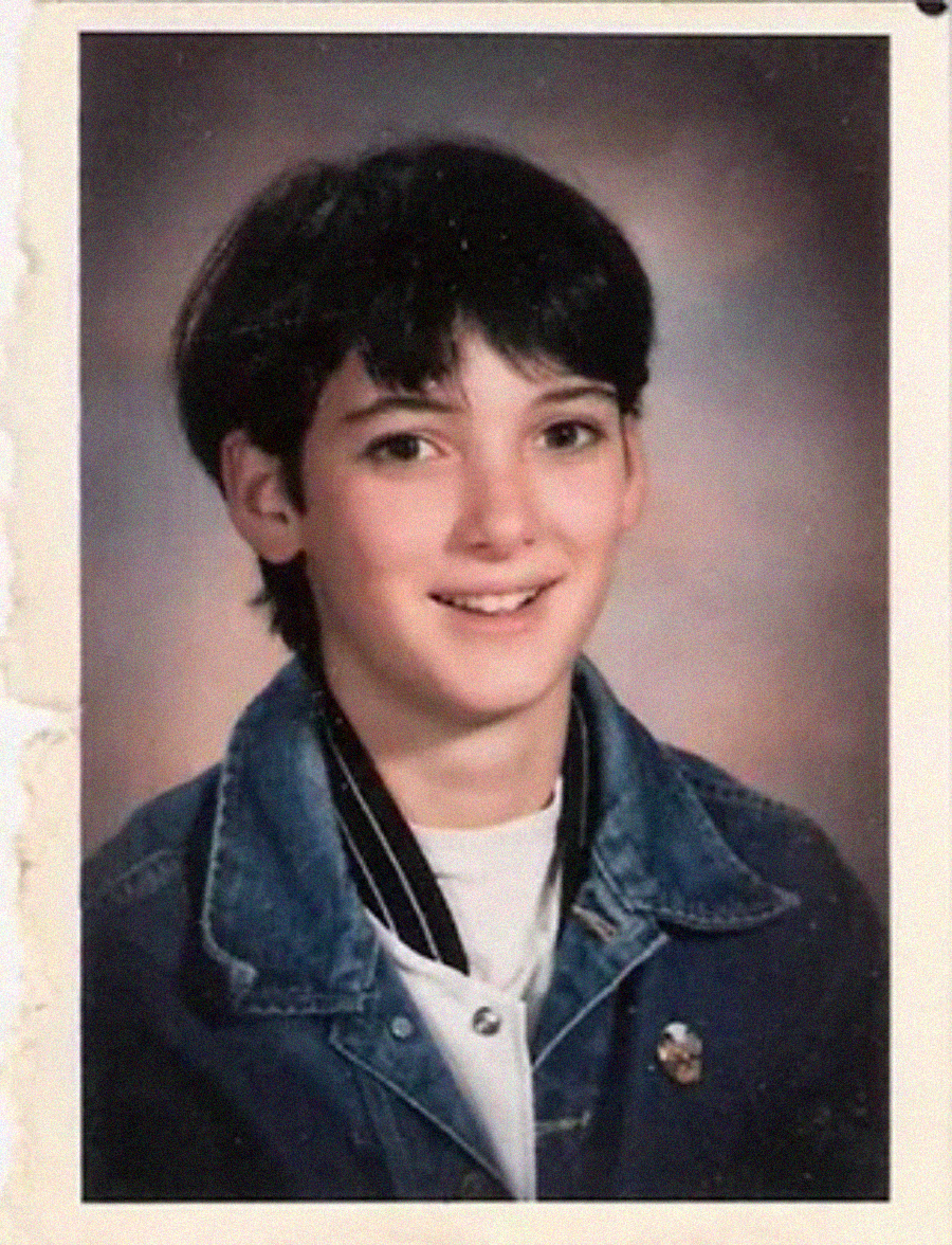 A young person with short dark hair smiles at the camera, wearing a denim jacket over a white shirt, against a blurred studio portrait background.