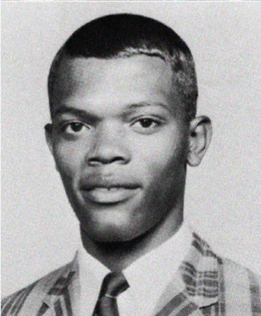 A young man in a plaid suit and tie poses for a formal black-and-white portrait, looking directly at the camera with a neutral expression.