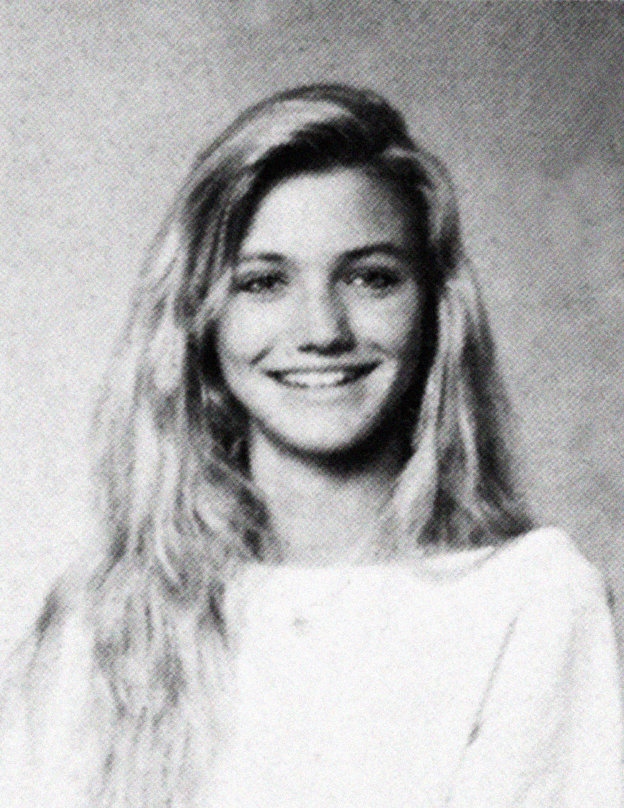 Black and white portrait of a young woman with long, wavy hair, smiling at the camera. She is wearing a light-colored top and is positioned in front of a plain backdrop.