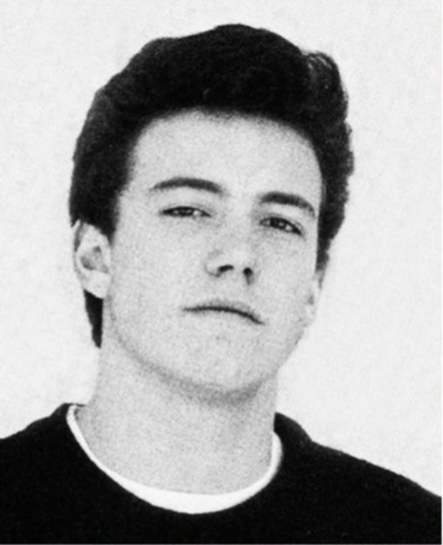 Black and white portrait of a young man with short, dark hair, wearing a dark crew-neck sweater over a light shirt, looking directly at the camera with a neutral expression.