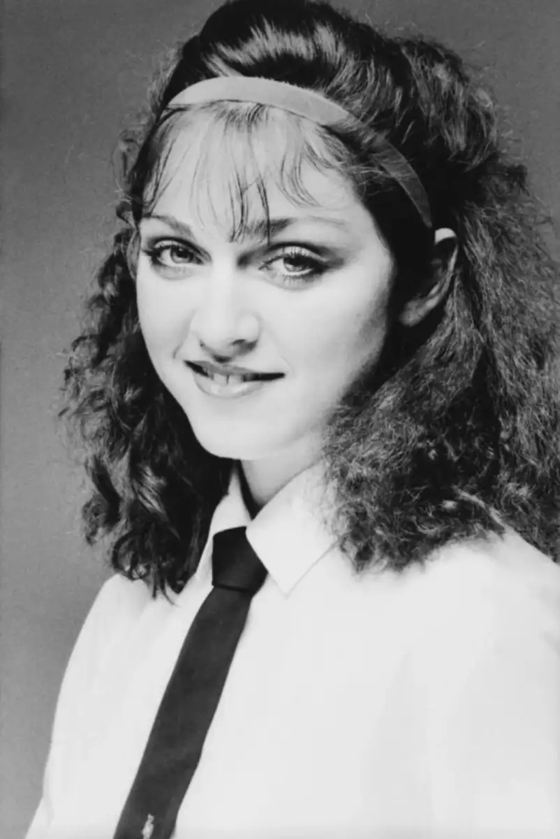 Young woman with wavy dark hair, wearing a white shirt, black tie, and a headband, smiles softly in a black-and-white portrait with a plain background.