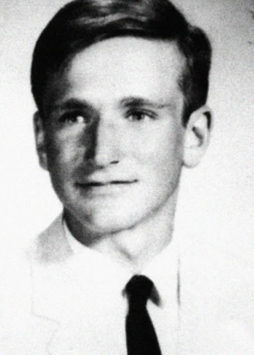 Black-and-white portrait of a young man with short hair, wearing a light-colored jacket, white shirt, and dark tie, looking slightly to the side and smiling softly.