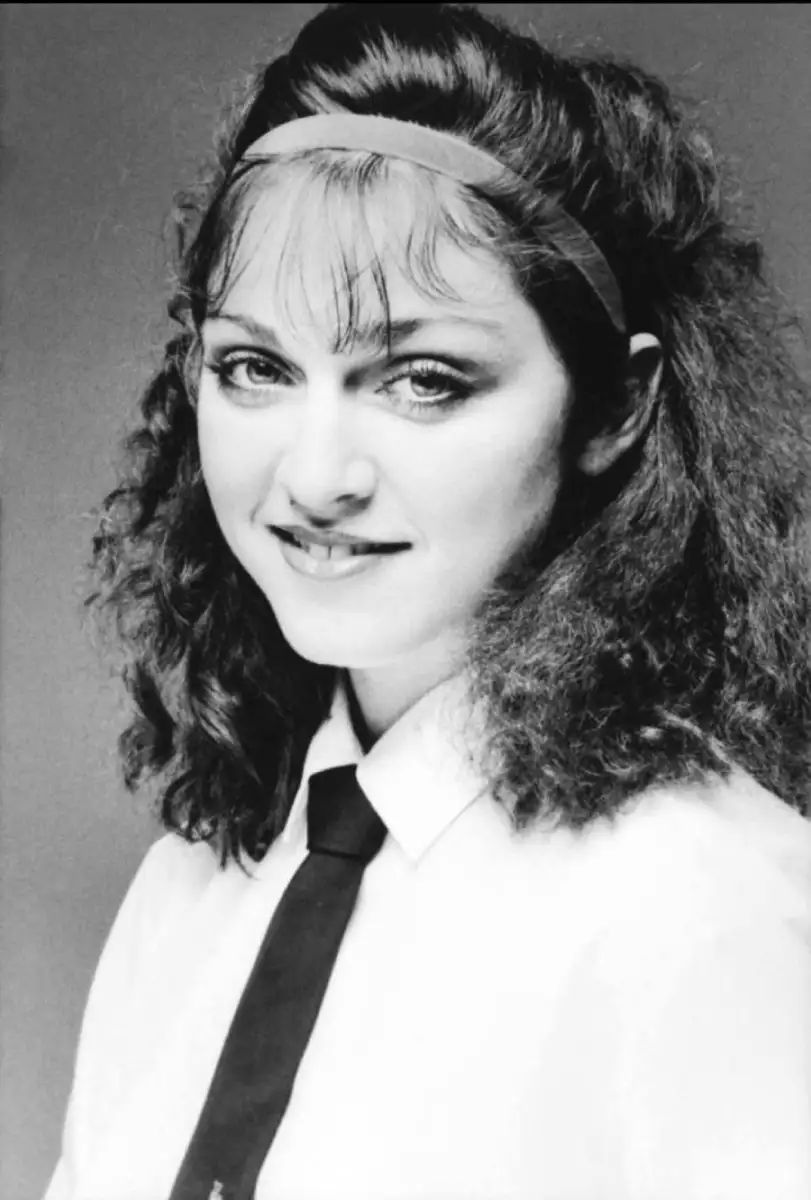 A young woman with curly dark hair and a headband smiles softly. She is wearing a white shirt and a black tie, facing the camera in this black-and-white portrait.