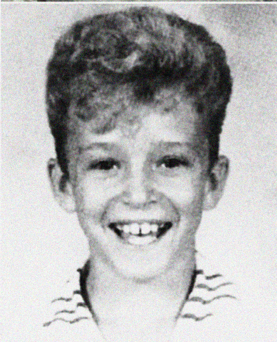 A black-and-white school portrait of a young boy with curly hair, smiling widely and showing his teeth. He is wearing a striped shirt and looking directly at the camera.