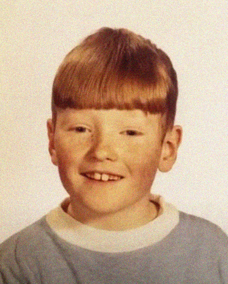 A young boy with light skin and straight reddish-brown hair cut in a bowl style smiles at the camera. He is wearing a light blue shirt with a white collar, posing in front of a plain light background.