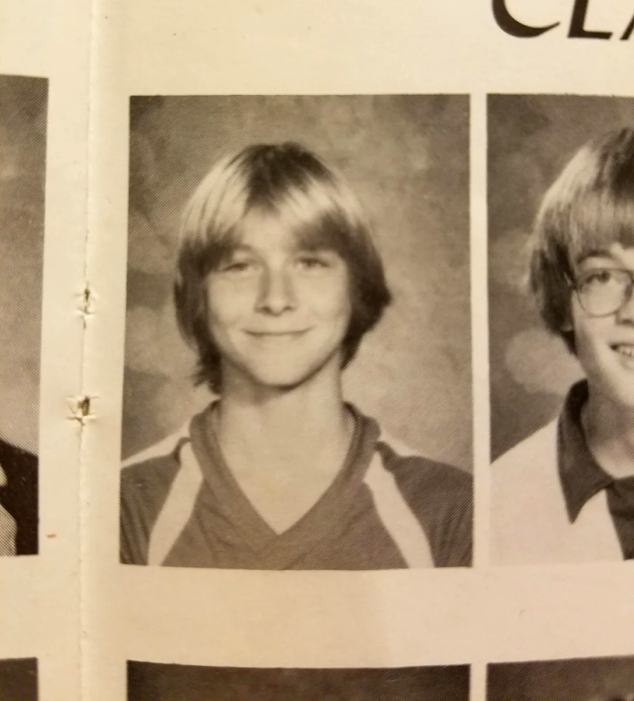 A black-and-white yearbook photo of a smiling young person with light-colored, medium-length hair wearing a sports jersey. The background is plain, and parts of two other portraits are visible on each side.