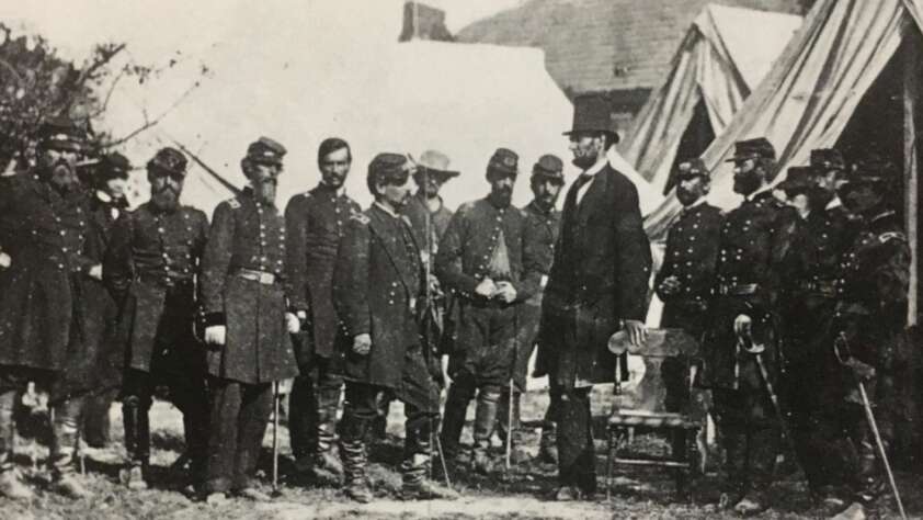 A historical black-and-white photo shows Abraham Lincoln standing among a group of Union soldiers and officers in uniform in front of military tents during the American Civil War.